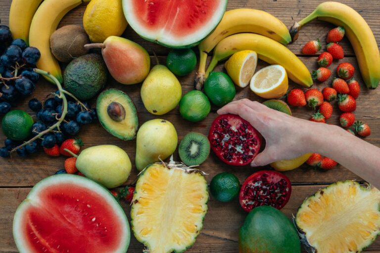 Colorful assortment of fresh fruits on wooden surface with a hand holding a pomegranate half.